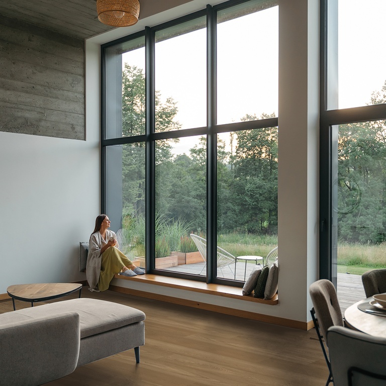 Bodiax Estrela 2BP370144 Toffee Oak PVC plank plak pvc in woonkamer met hoge ramen en uitzicht op het groen sfeerfoto